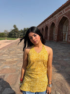 A woman wearing a yellow and white patterned halter top stands outdoors in front of a historic red sandstone archway building under a clear blue sky.