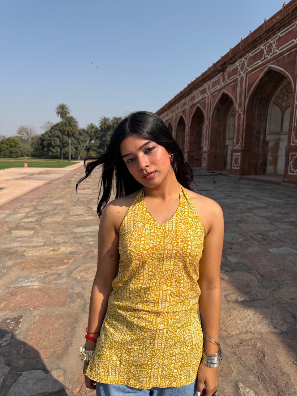 A woman wearing a yellow and white patterned halter top stands outdoors in front of a historic red sandstone archway building under a clear blue sky.