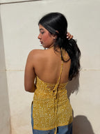 A woman shown from the back wearing a yellow and white patterned halter top with lace-up detailing, paired with blue jeans, standing against a light beige wall.